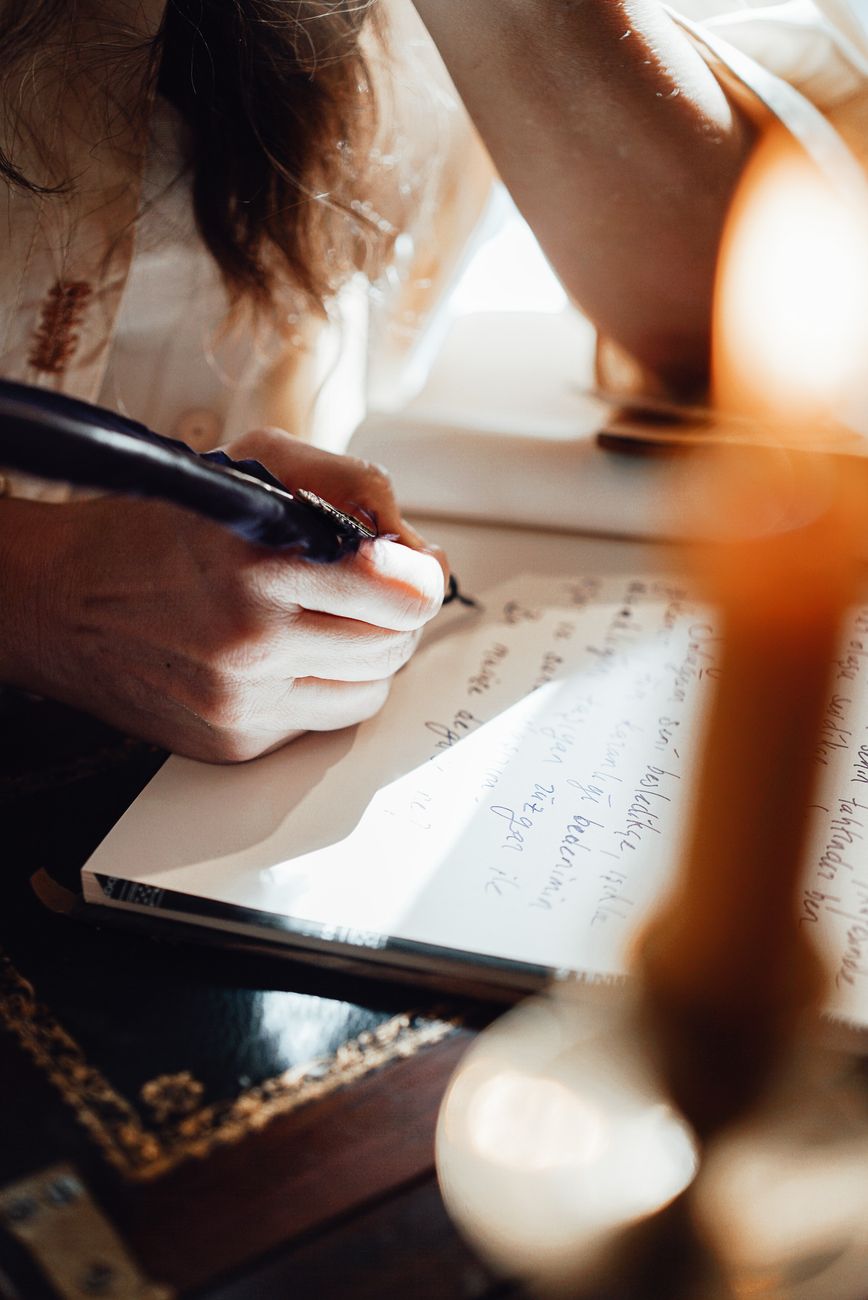 A close-up of a person's hand writing with a pen on a notebook, illuminated by soft light from a nearby candle.